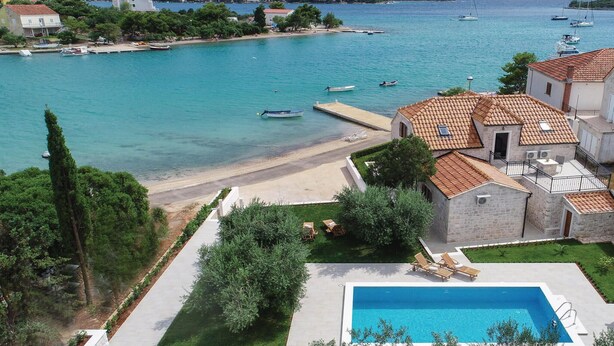 A coastal villa with a swimming pool, a beach, and boats in the background.