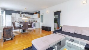 A modern living room with a sofa, ottoman, and glass coffee table. A kitchen with white cabinets and a dining area with a table and chairs is visible in the background.