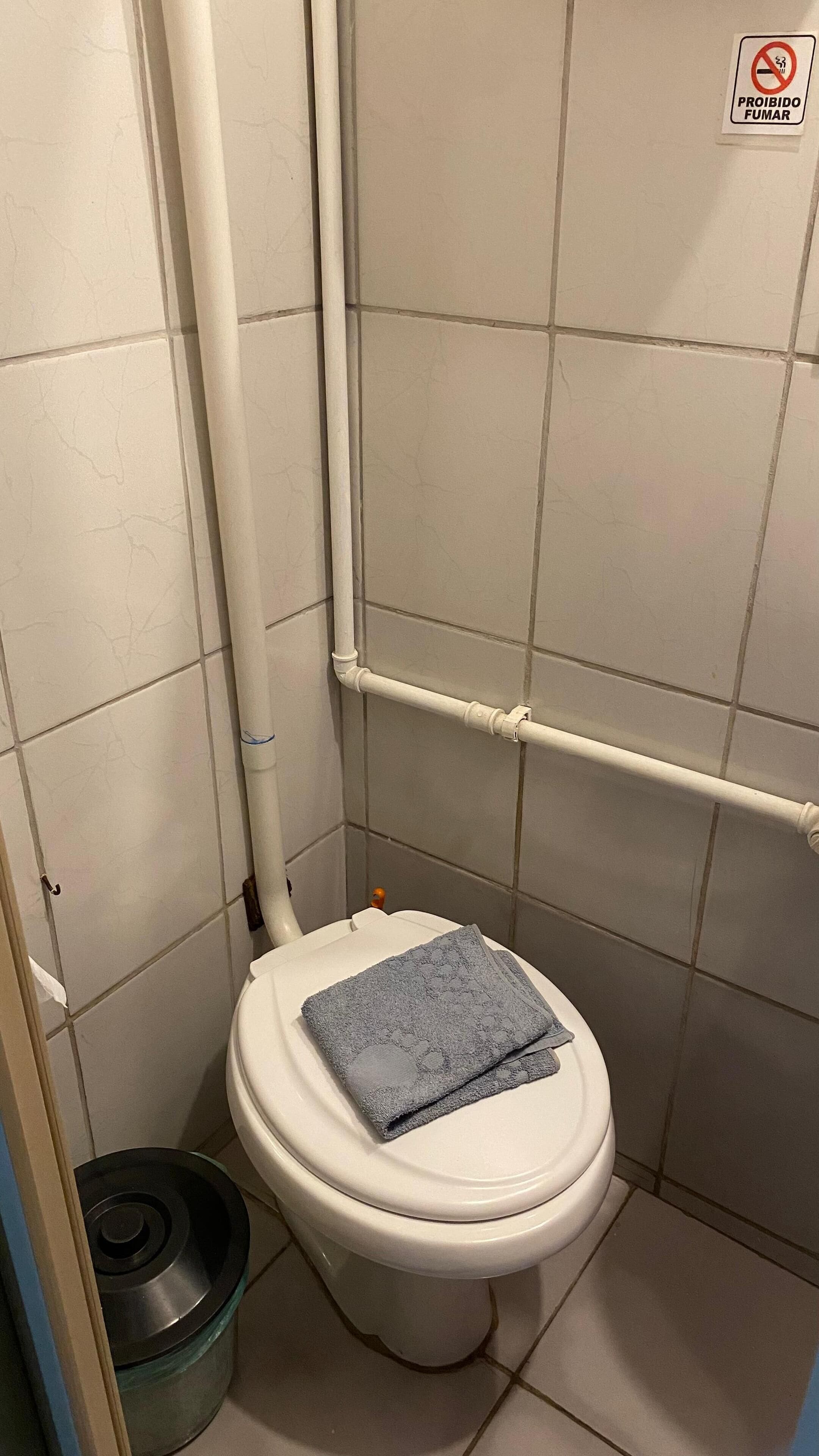 A bathroom with a toilet, a white pipe, and a 'PROIBIDO FUMAR' sign.