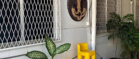 Un patio con baldosas, una planta en maceta, una silla amarilla y azul, y un tapiz decorativo.