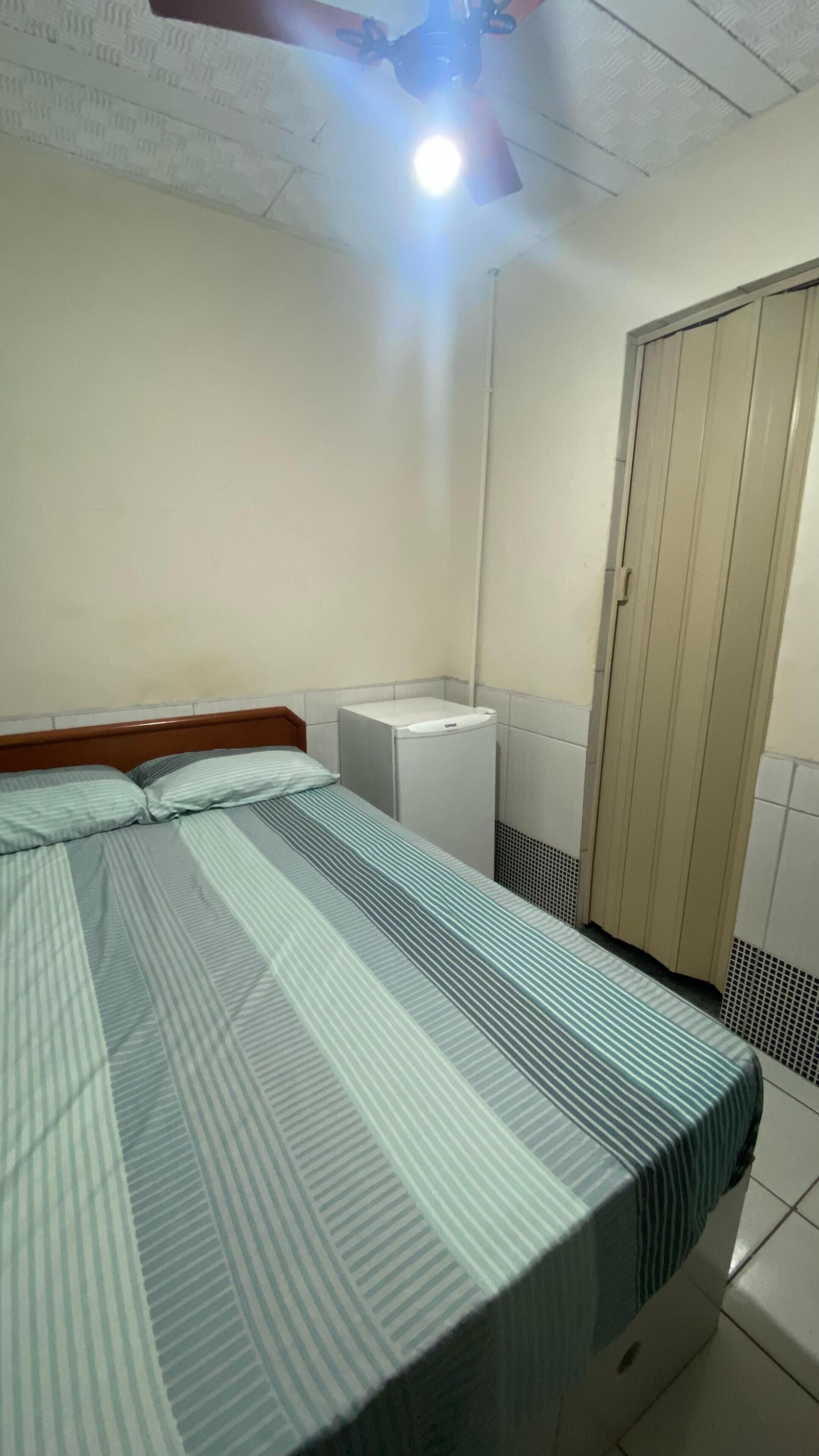Una habitación pequeña y sencilla con una cama, un ventilador de techo, un calentador blanco y una puerta.
