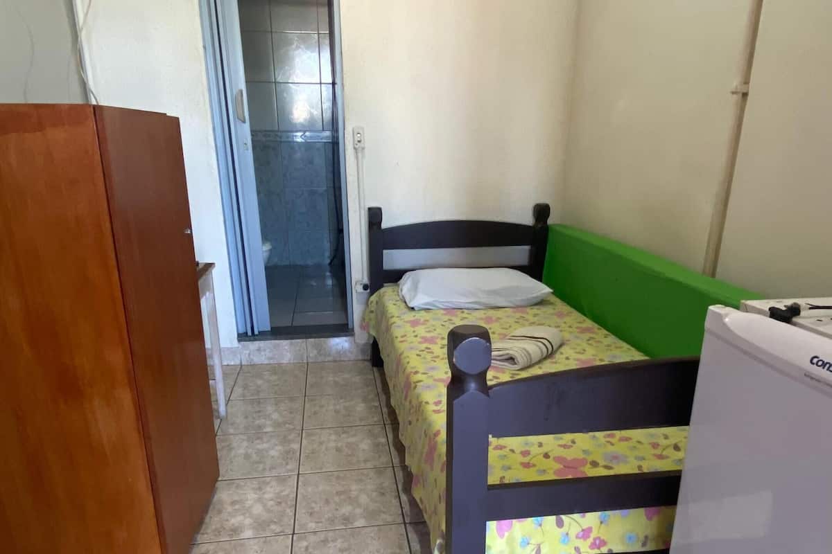 A small, compact room with a bed, a wooden wardrobe, a small table, and a white refrigerator.