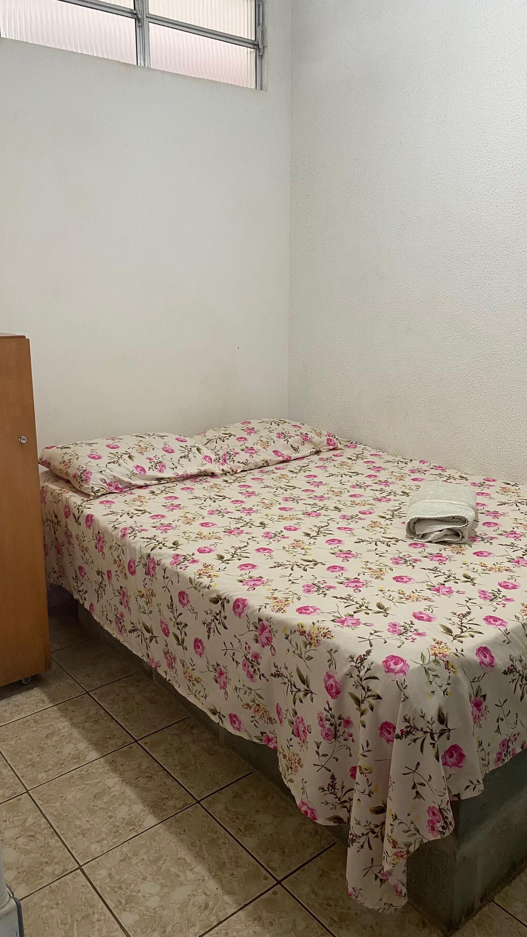 Una habitación pequeña con cama individual, colcha estampada, puerta de madera y suelo de baldosas.
