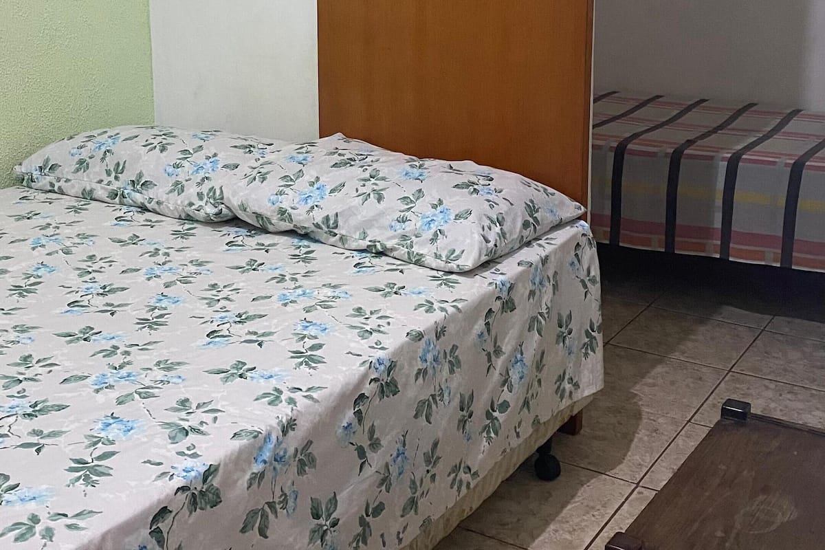 A bedroom with a bed, a wooden wardrobe, a small table, and a window with blinds.