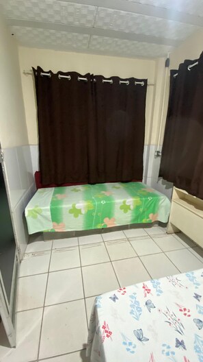 A small, enclosed room with a bed covered in a green floral bedspread, a brown curtain, and a white tiled floor. - Pousada e Alojamento do Engenho de Itaguai (Itaguai)