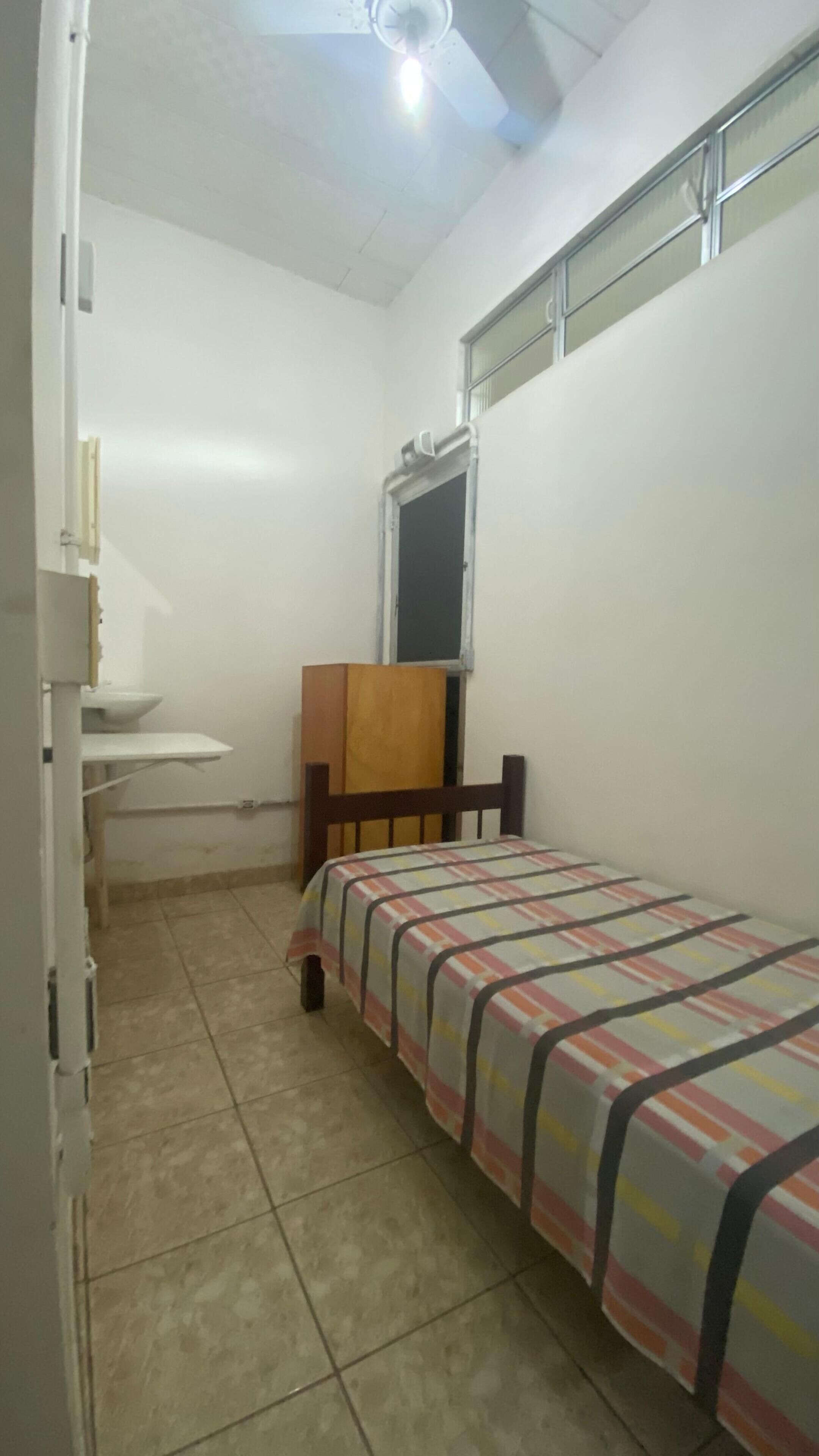 Ein kleines, sauberes Zimmer mit einem Einzelbett, einem Holzstuhl, einem kleinen Tisch und einem Fenster mit Metallrahmen.
