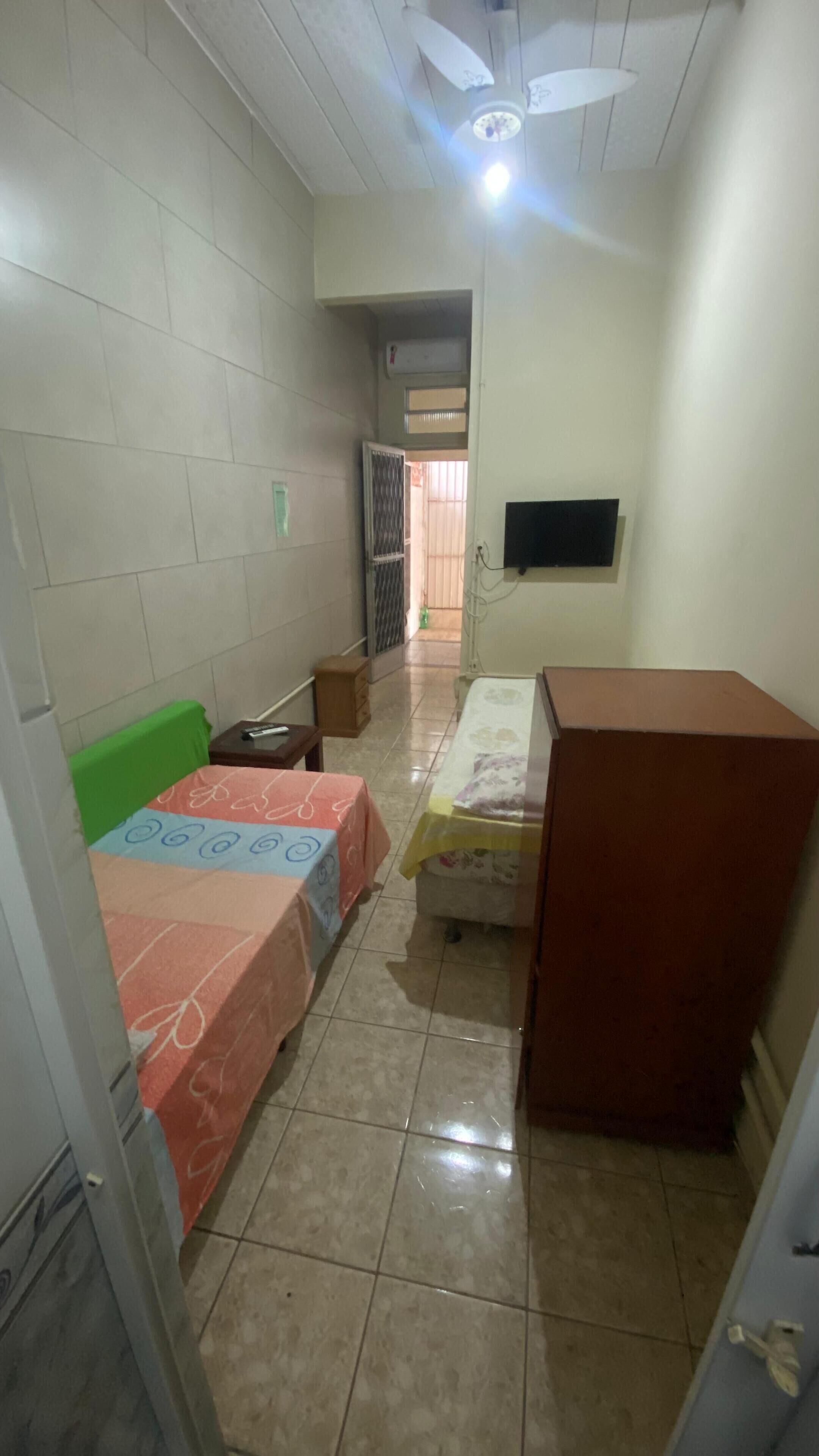 A small, tiled room with two beds, a television mounted on the wall, and a ceiling fan.