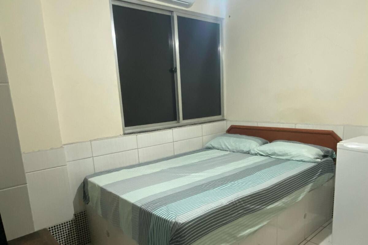 A bedroom with a bed, a ceiling fan, an air conditioner, a window, and a small table with a phone.