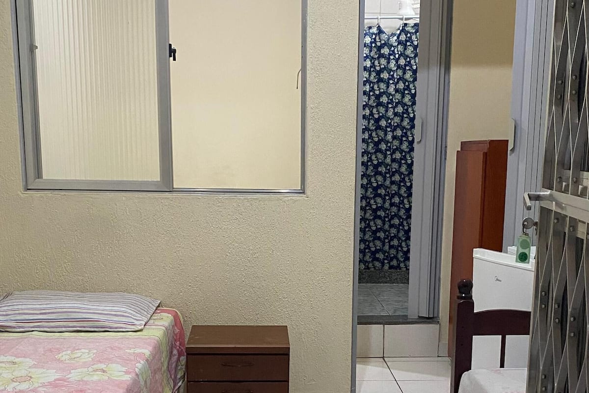 A bedroom with a bed, a small wooden chest, a window with blinds, a door leading to another room, and a metal gate.