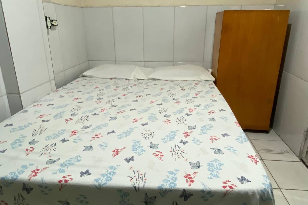 A small, single-bed room with a floral-patterned bedspread, white walls, and a wooden wardrobe.