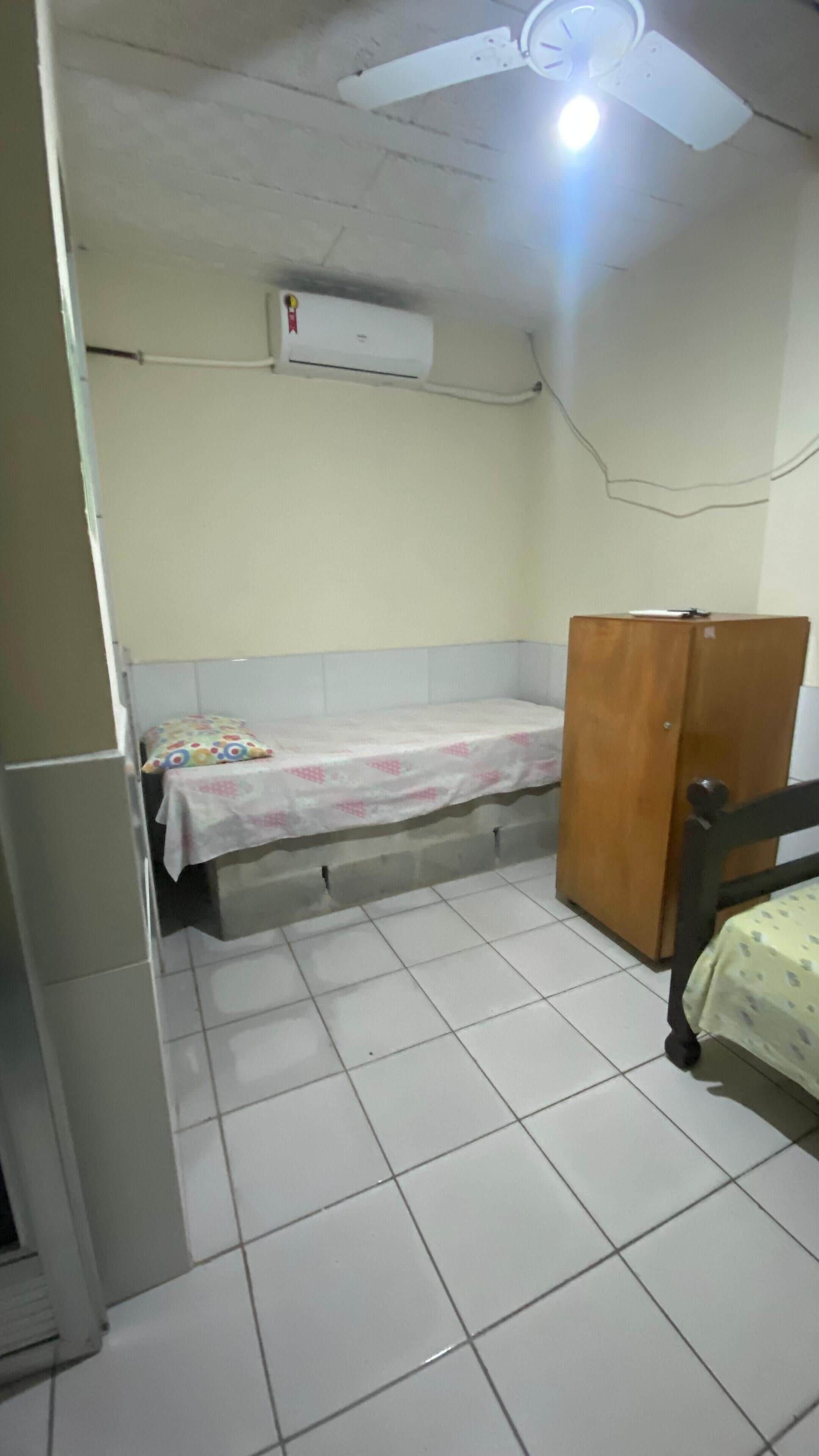 Una habitación pequeña con baldosas, una cama, un armario de madera y un aire acondicionado en la pared.