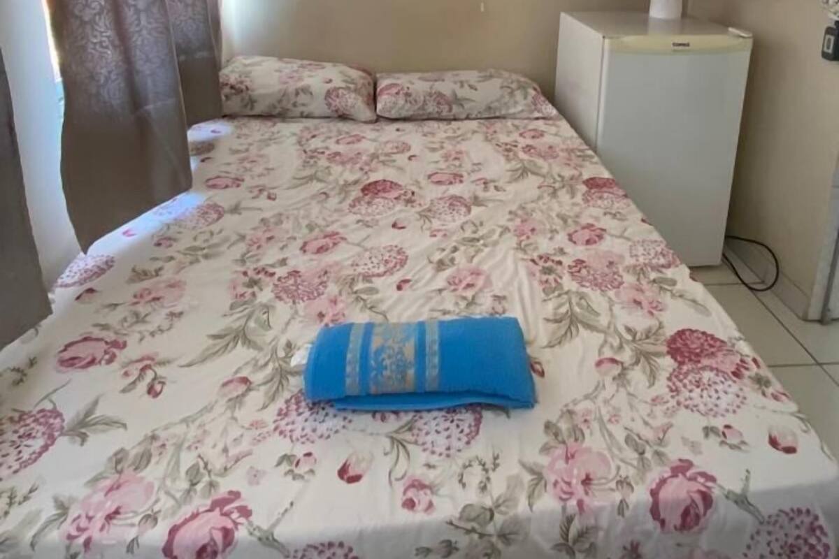 A bedroom with a bed, floral bedspread, a blue towel, a wooden wardrobe, a window, and a ceiling fan.