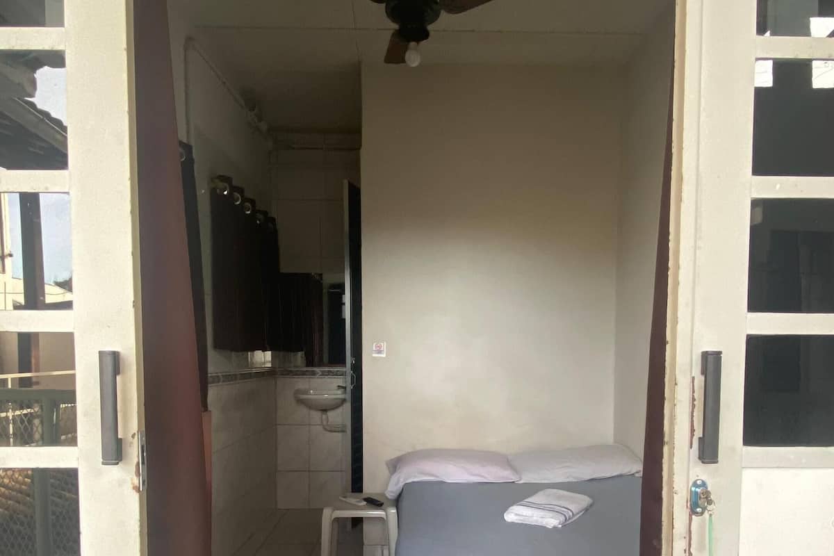 A narrow room with a bed, a ceiling fan, and a small stool.