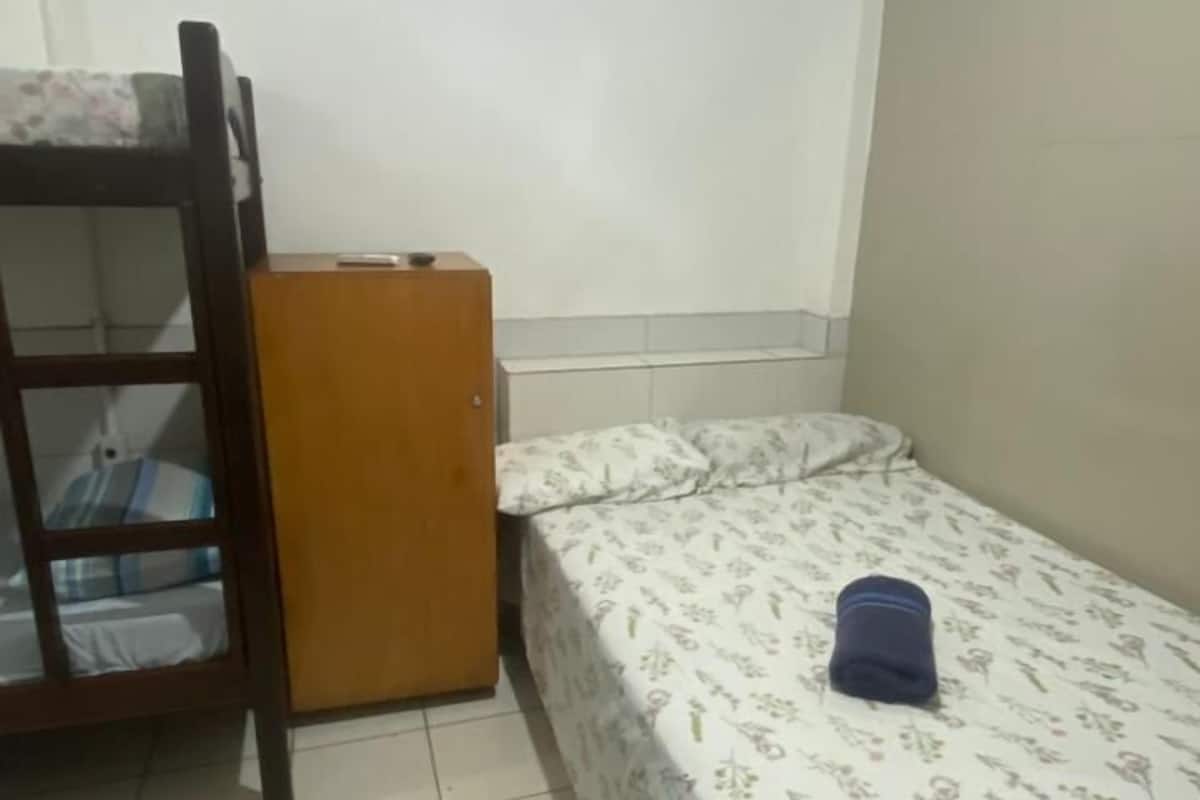 A small, sparsely furnished room with a bunk bed, a small wooden cabinet, and a single bed with a floral bedspread.