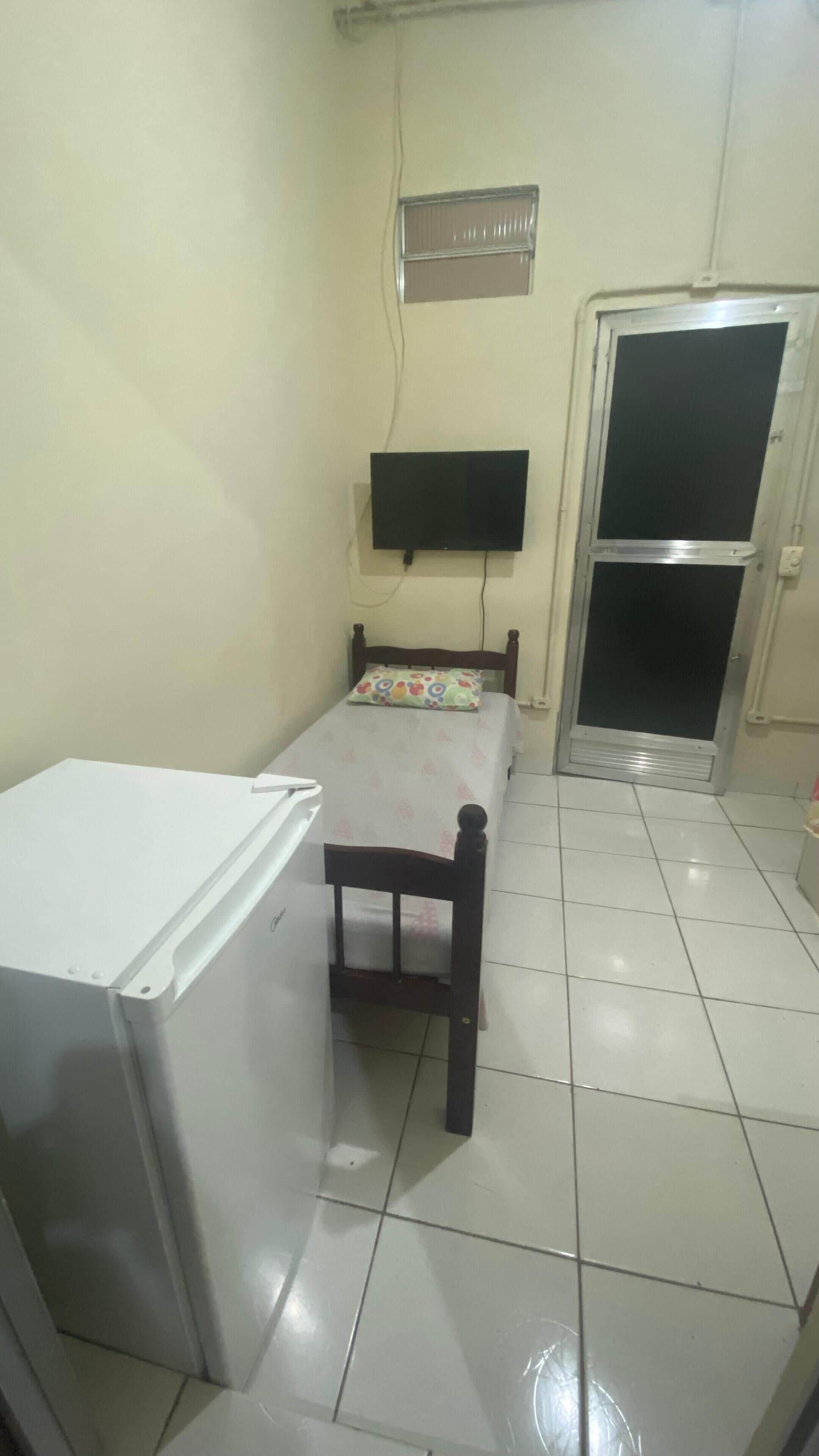 A small, compact room with a bed, a white refrigerator, a TV mounted on the wall, and a door with a window.
