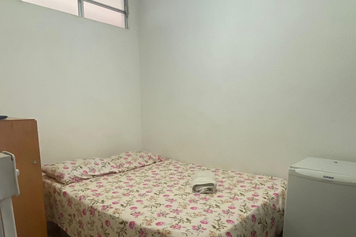 A small, single-bed room with a floral bedspread, a ceiling fan, a window, and a refrigerator.