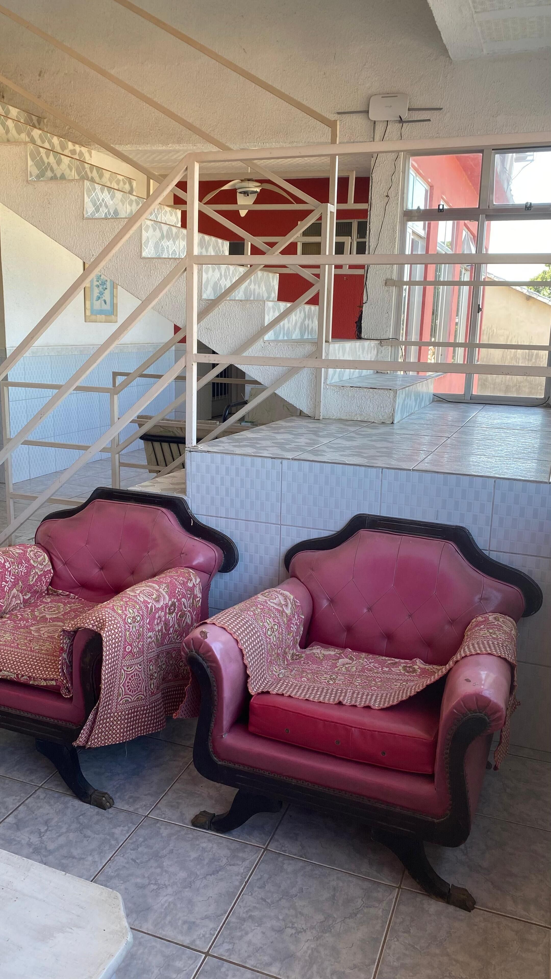 A room with two red upholstered chairs, a tiled floor, a staircase with a red wall, and a ceiling fan.