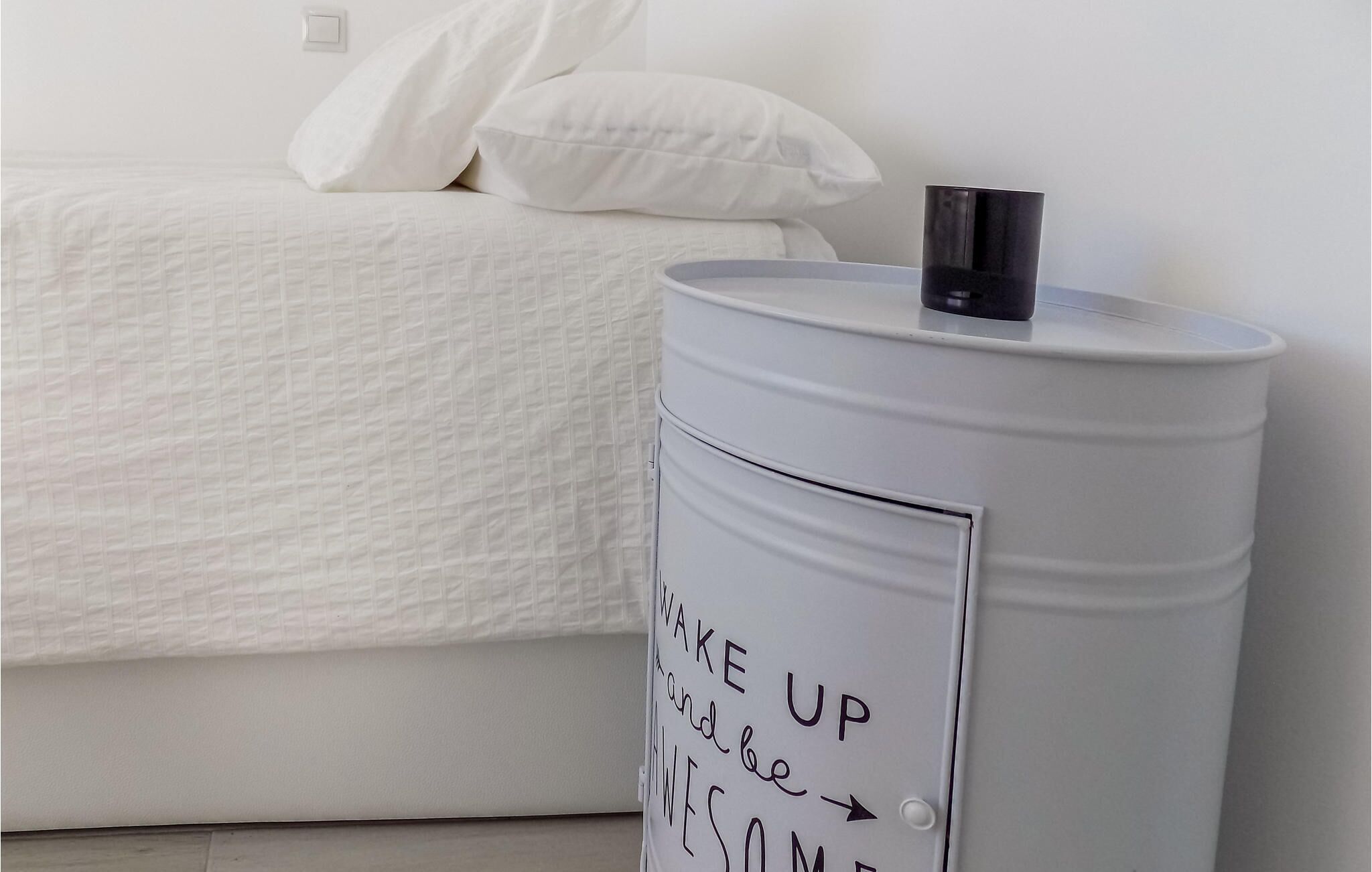 Una cama con ropa blanca y almohadas, una mesita de noche con un objeto negro encima, y un cartel sobre la mesa que dice "WAKE UP and be AWESOME".