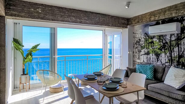 Un salón moderno con balcón vistas al mar, que incluye una mesa de comedor con sillas, un sofá y un ventanal grande.