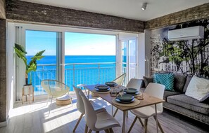 Ein modernes Wohnzimmer mit Balkon und Meerblick, ausgestattet mit einem Esstisch und Stühlen, einer Couch und einem großen Fenster.