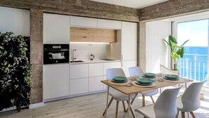 Una cocina moderna con armarios blancos y un estante superior de madera, un comedor con mesa para cuatro personas, y un balcón con vista al mar.
