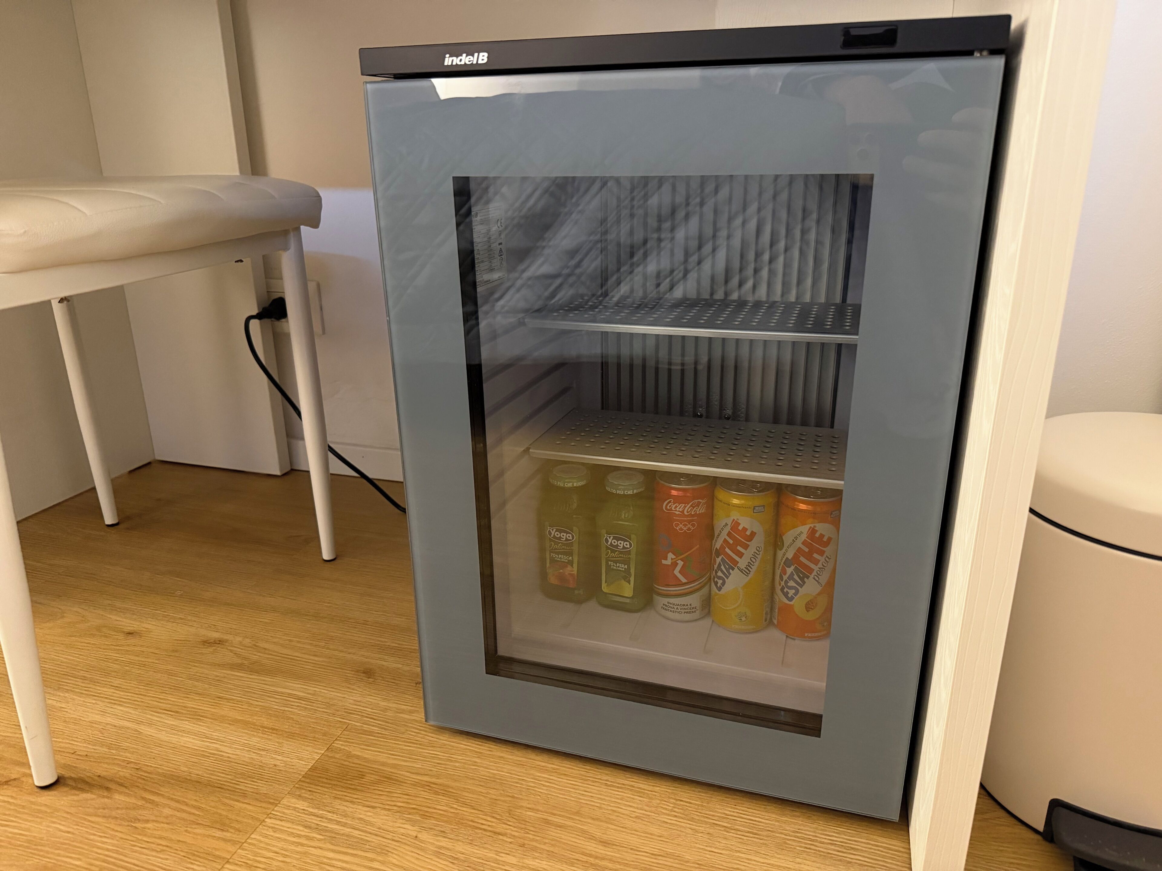 Un refrigerador pequeño y moderno con puerta de vidrio, que contiene bebidas y un envase de yogur.