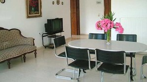 A dining area with a round table, six chairs, a television on a stand, a sofa, and a painting on the wall. - Dimitra Apartments and Studios Gouvia (Corfu)