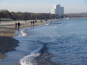Une plage où des gens se promènent le long du rivage, une jetée qui s’avance dans l’eau et, au loin, un grand immeuble.
