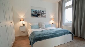 A bedroom with a bed, two bedside lamps, a nightstand, a window with curtains, and a wall-mounted picture. - Holiday apartment (6) Zudar - Apartment house Johanneshorst (Sellin)