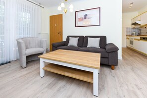 Un salon moderne avec un canapé gris, un fauteuil gris clair et une table basse en bois. On peut voir une porte en bois et un tableau abstrait encadré au mur.