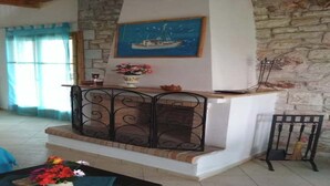 Un salón con pared de piedra, un cuadro de un barco, chimenea con barandilla de metal y una mesa con flores.