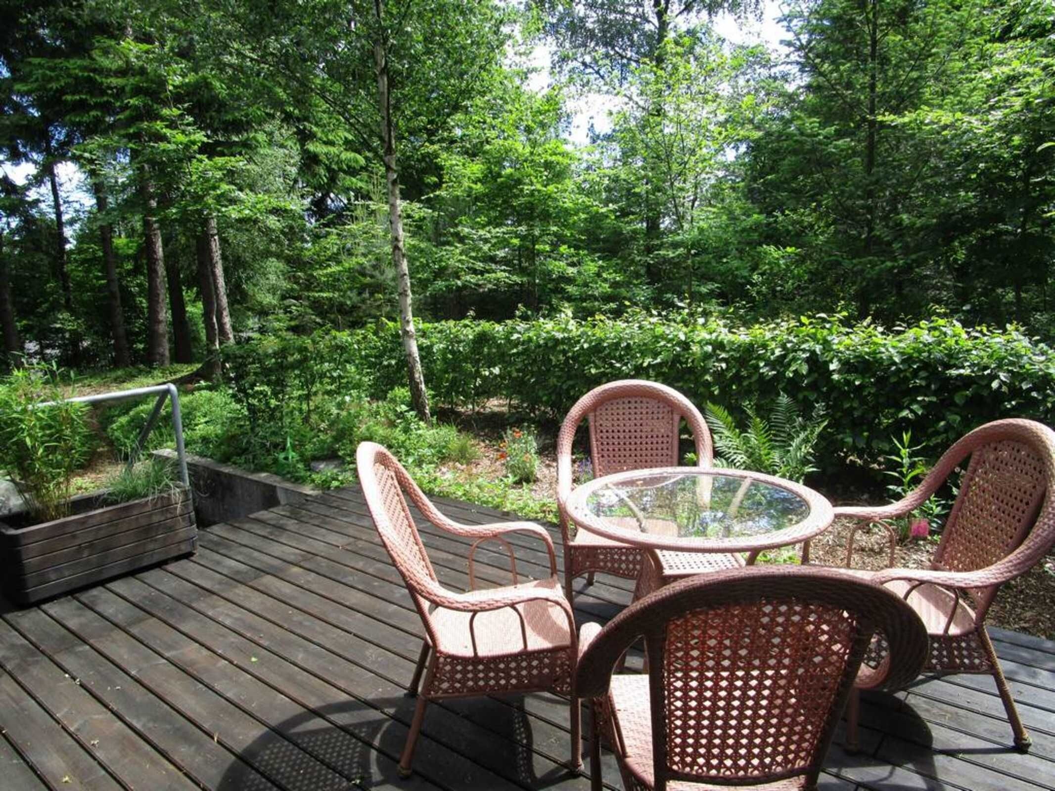 Eine Holzterrasse mit Korbstühlen und einem Glastisch, umgeben von üppiger Bepflanzung.