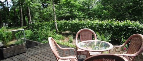 Eine Holzterrasse mit Korbstühlen und einem Glastisch, umgeben von üppiger Bepflanzung.