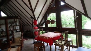 A dining area with a table covered in a red tablecloth, surrounded by wooden chairs, under a sloped ceiling with exposed beams.