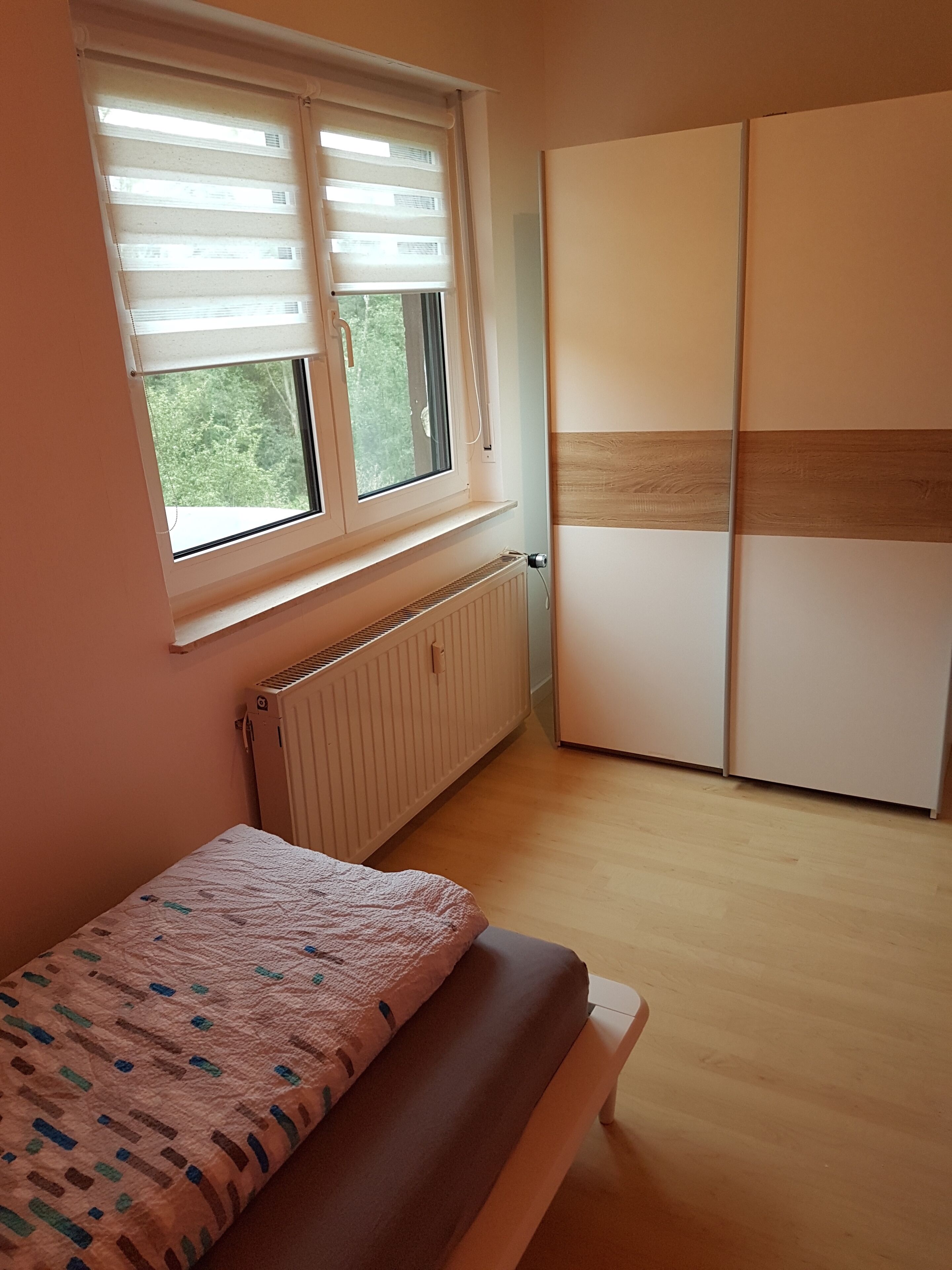 A bedroom with a bed, a radiator, a window with blinds, and a wardrobe.