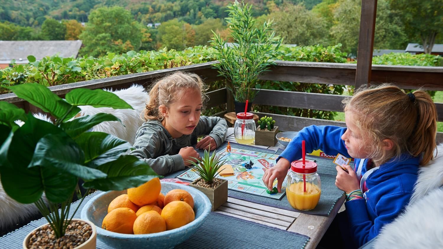 Eine Holzterrasse mit einem Tisch für zwei Personen, mit einer Schale Orangen, einem Blumentopf und einem Getränk mit Trinkhalm.