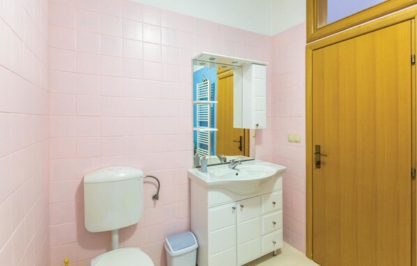 Un bany amb parets revestides de rajoles roses, un lavabo blanc, un mirall, un vàter i una porta de fusta.