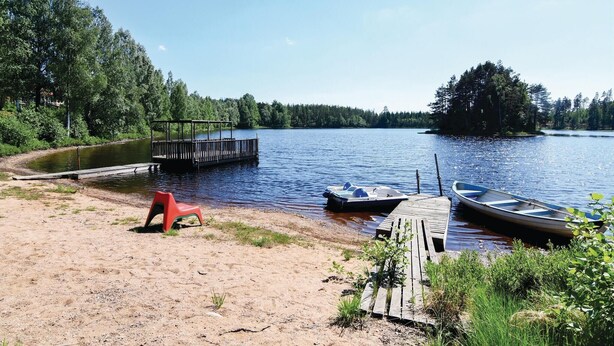 A lakeside setting with a wooden dock, a small boat, and a red chair on the sandy shore. - Gorgeous home in Ambjörnarp with WiFi (Ambjörnarp)