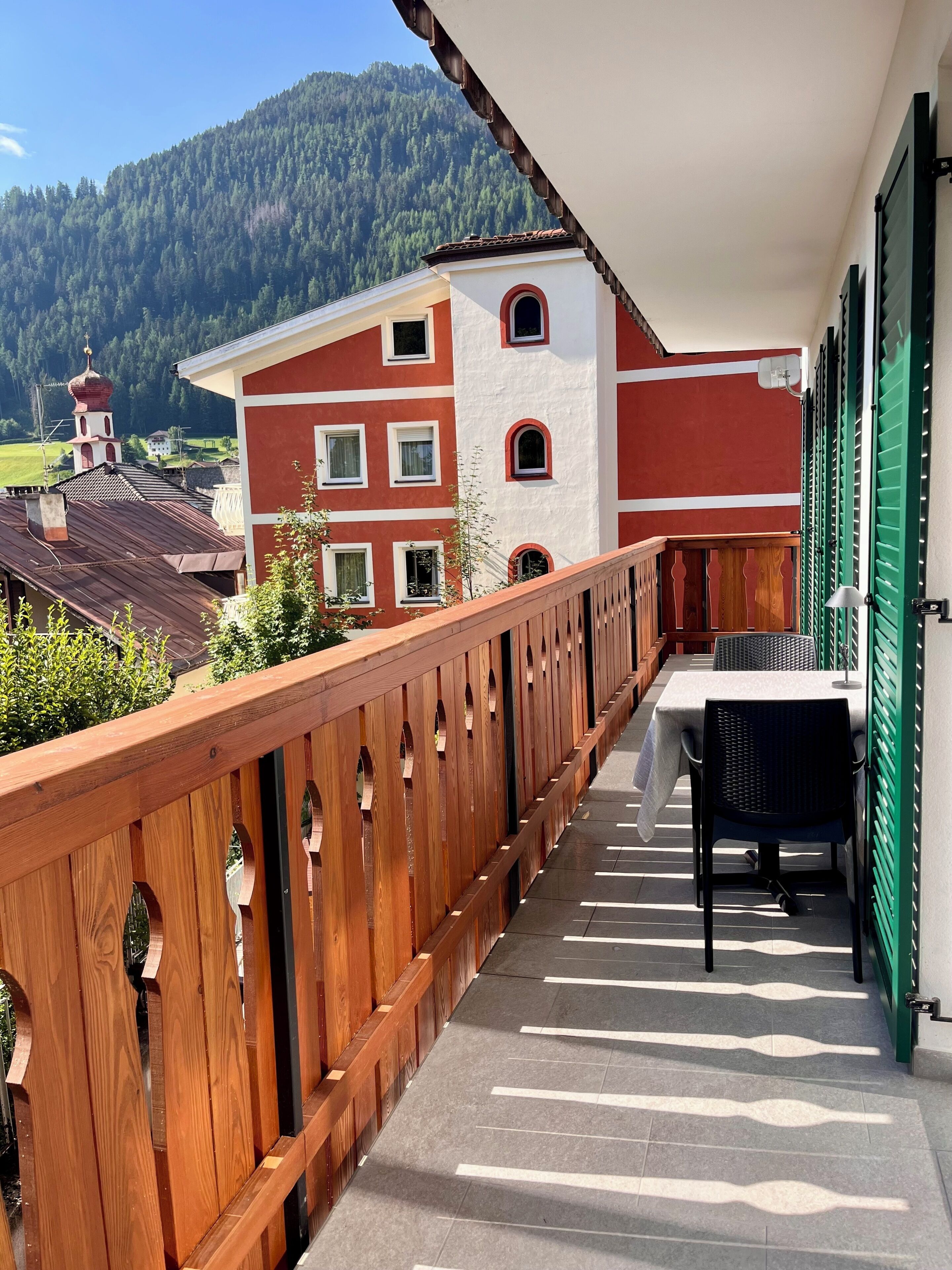 Un balcon doté d’une balustrade en bois, d’une table et de chaises, offrant une vue sur un paysage montagneux avec un bâtiment en arrière-plan.