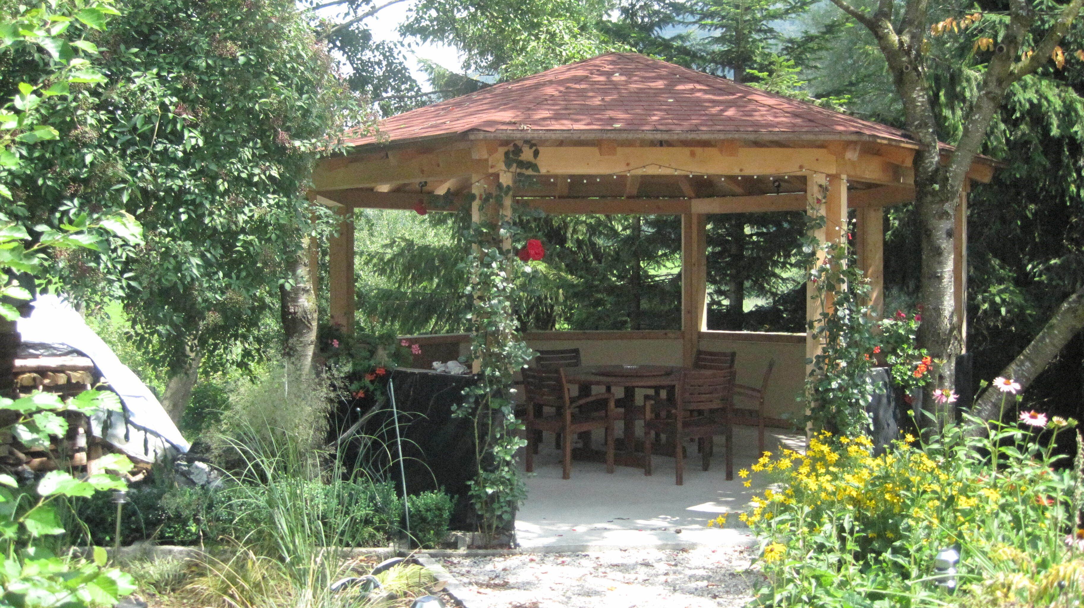 Um gazebo com telhado de cerâmica, rodeado por vegetação e uma mesa de jantar com cadeiras.