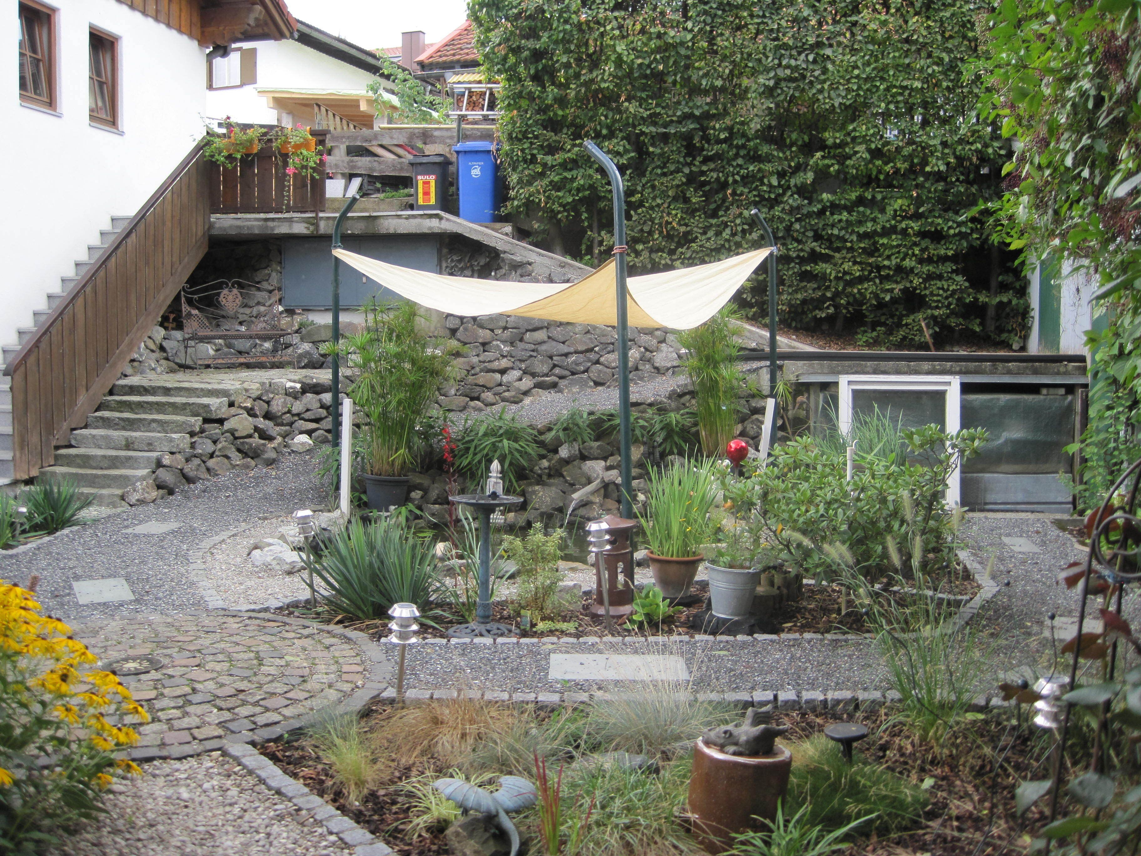 Un jardín con un sendero de piedra, plantas en macetas, una pequeña fuente y una escalera de madera que lleva a una casa.