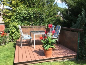 Une terrasse en bois avec une table et deux chaises, entourée de verdure et d’une clôture.