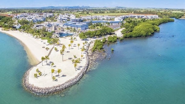 Vist panoràmic d'un complex turístic costaner amb una platja de sorra, palmeres i una massa d'aigua.