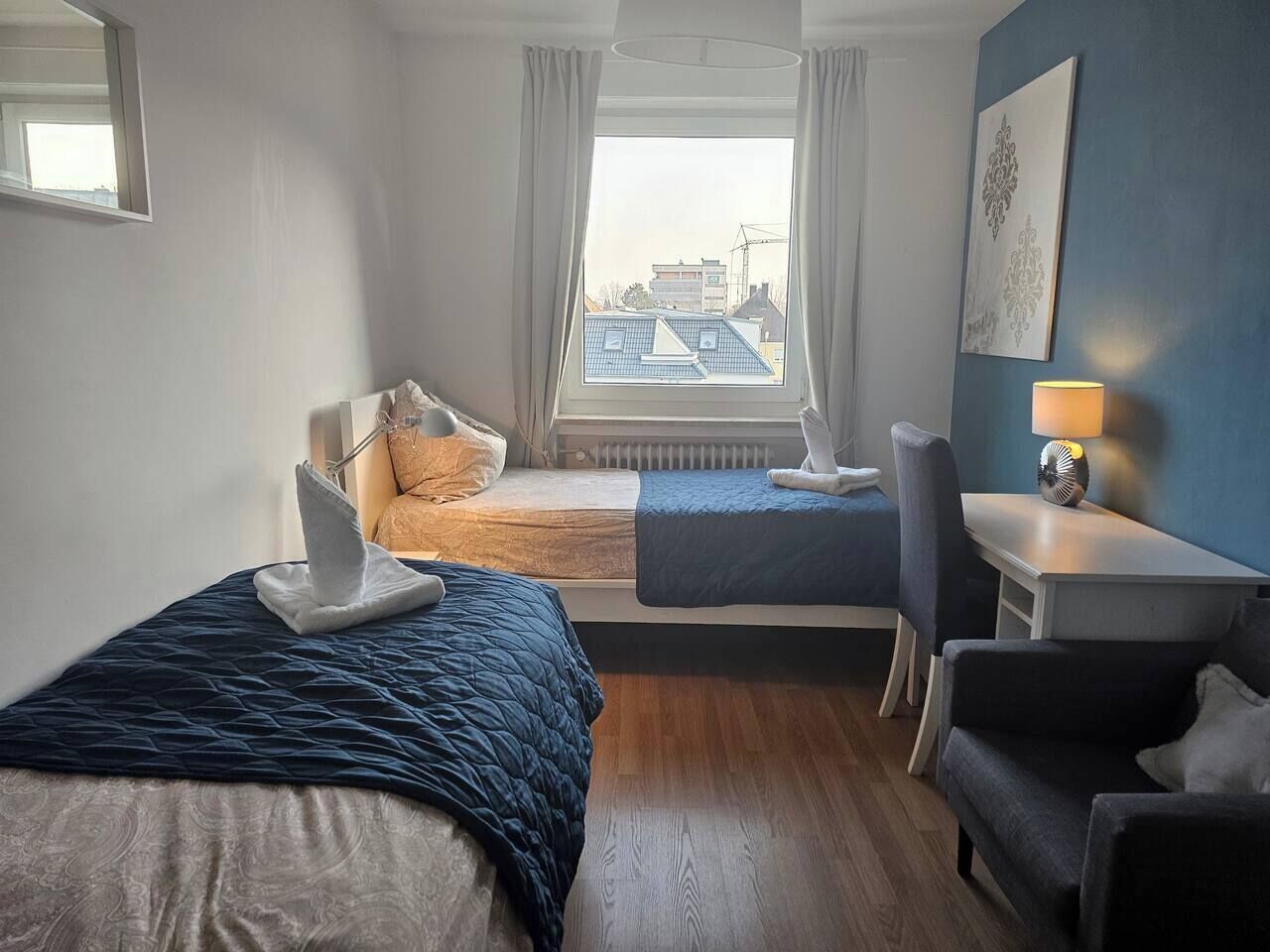 Habitación de hotel con dos camas, un escritorio, una silla y vistas a edificios a través de la ventana.