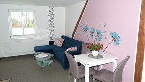 Una sala de estar con un sofá azul, una mesa de centro de vidrio, una mesa blanca con dos sillas y un jarrón con flores rojas.
