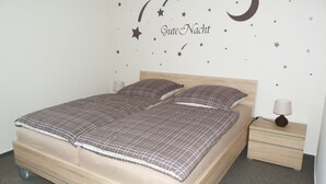 Una cama bien hecha con colcha de cuadros y dos almohadas, una mesita de noche con lámpara, y una pared decorada con estrellas y lunas.