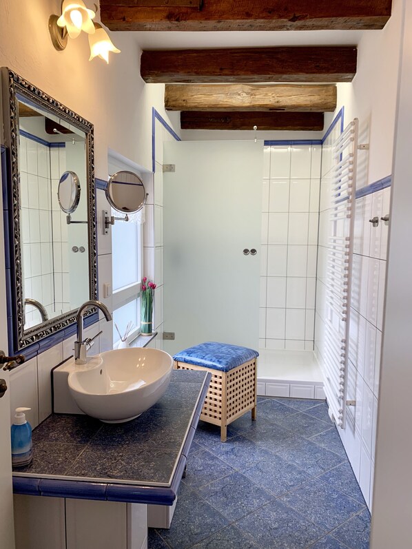 Une salle de bain avec un lavabo blanc, deux miroirs, un tabouret en bois et un espace douche.