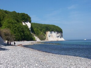 Une plage de galets bordée d’une falaise blanche et de verdure, un bateau sur l’eau et des personnes qui se promènent.