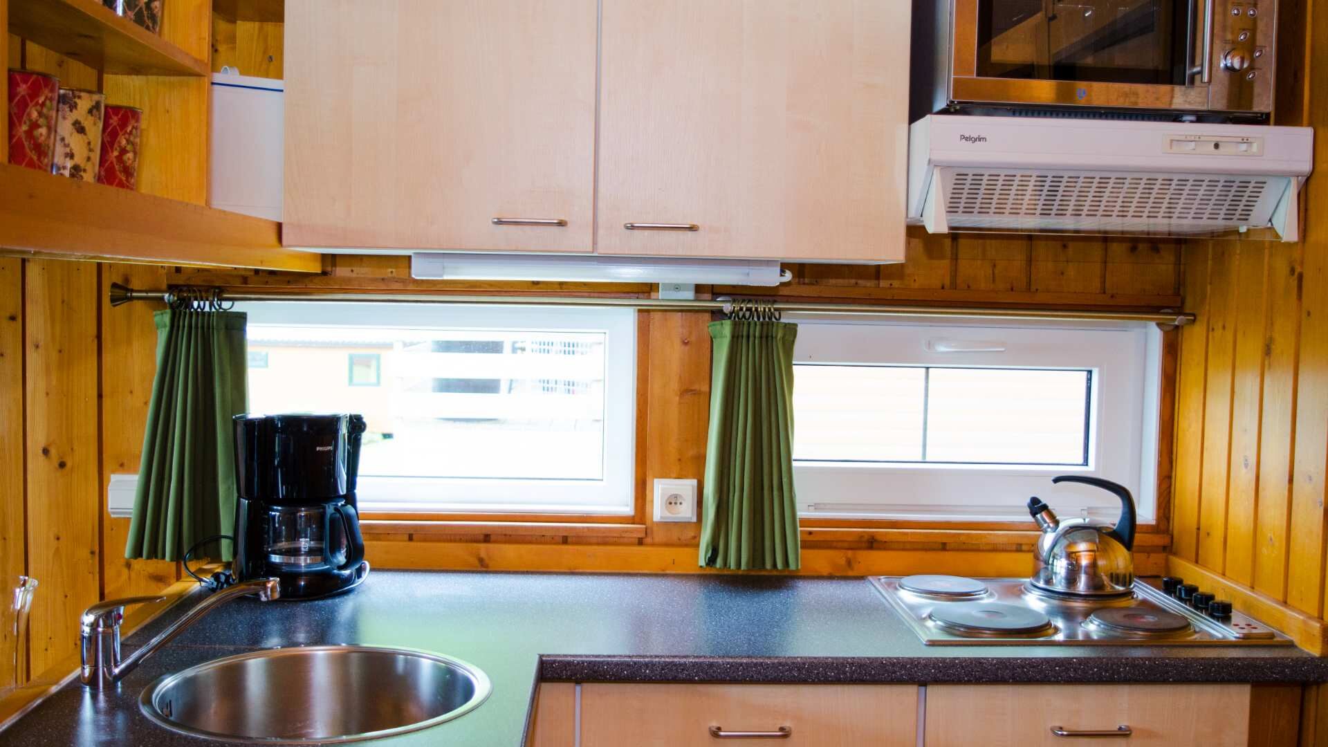 Een kleine keuken met houten kastjes, een spoelbak, een koffiezetapparaat, een waterkoker en een raam met gordijnen.