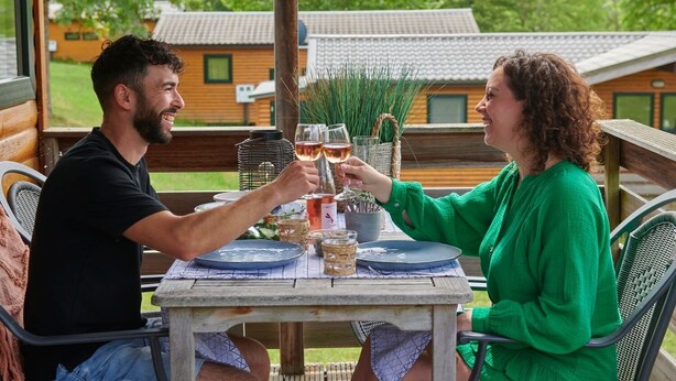 Deux personnes trinquent autour d’une table en bois, des verres de vin à la main, entourées de verdure et avec une vue sur des maisons.